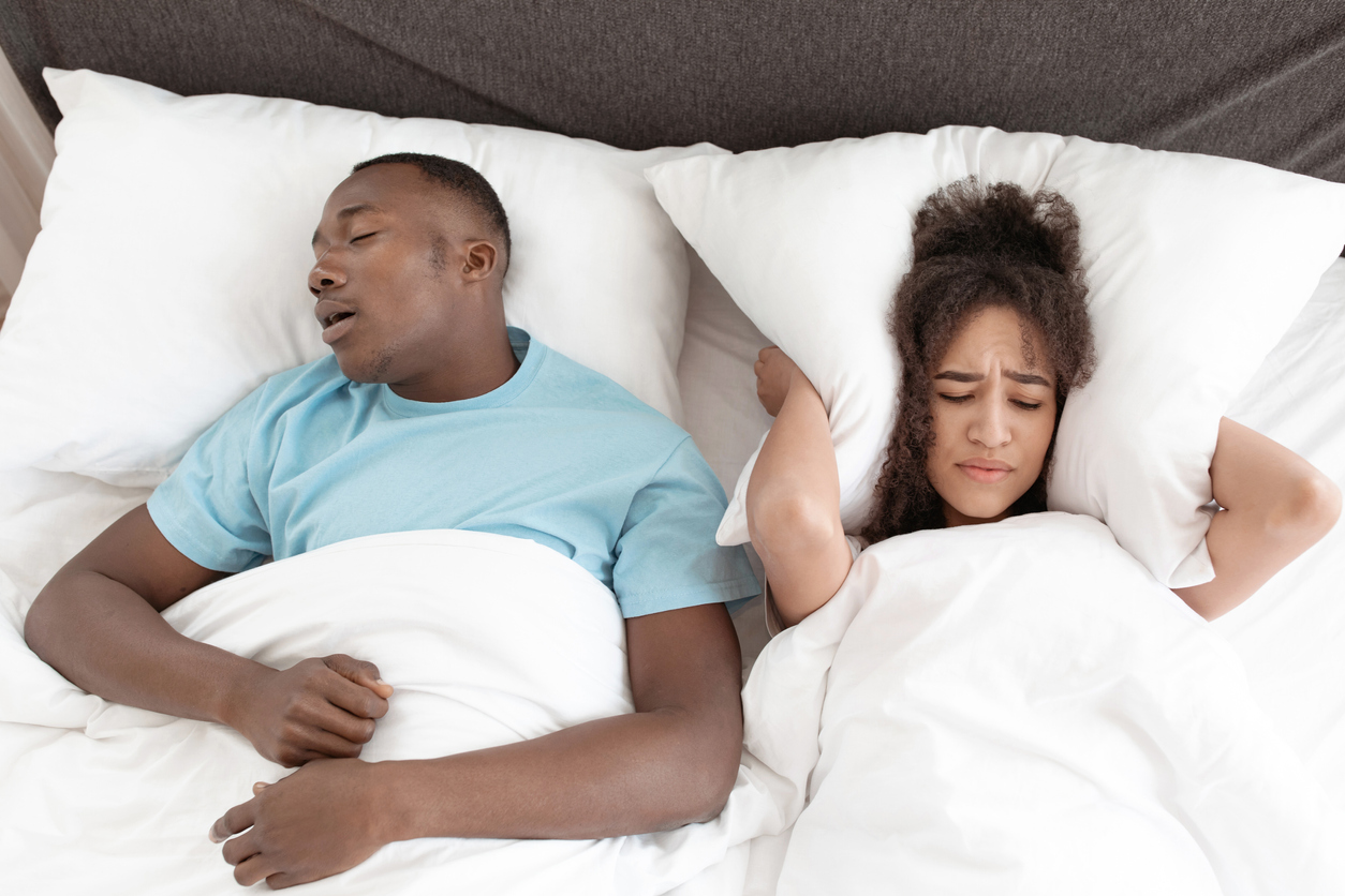 A couple lying in bed with a male snoring loudly and the woman covering her ears with a pillow. 