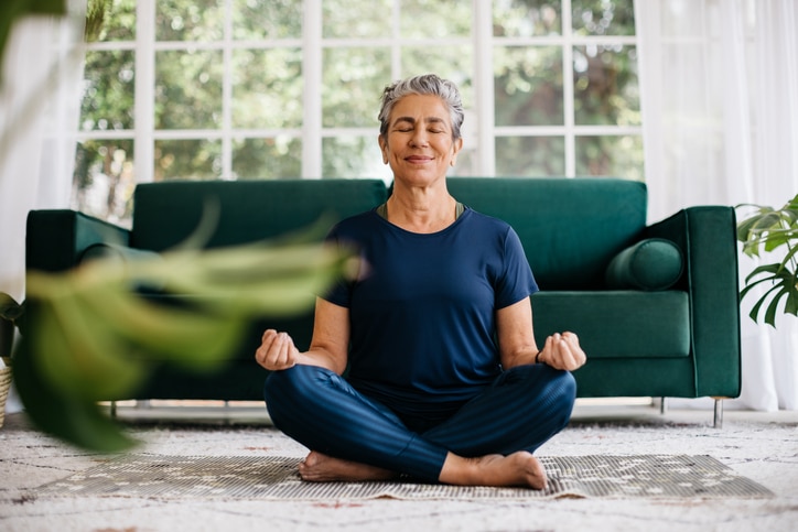 An Older Woman In A Blue Jumpsuit Meditates On A Grey Rug In Front Of A Green Sofa In Front Of A Window With White Curtains.