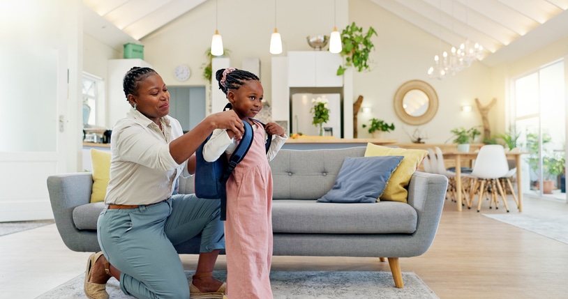 A Mother In A White Shirt And Jeans Puts A Blue Backpack On Her Daughter, Who Is Wearing A Pink Jumpsuit, In Front Of A Grey Sofa.