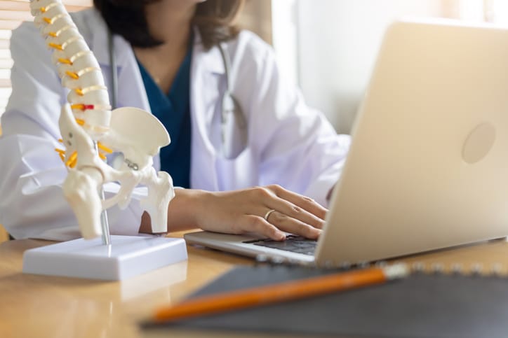 Female bone and joint health doctor types on a laptop with a human skeleton anatomy model next to the computer on her desk.