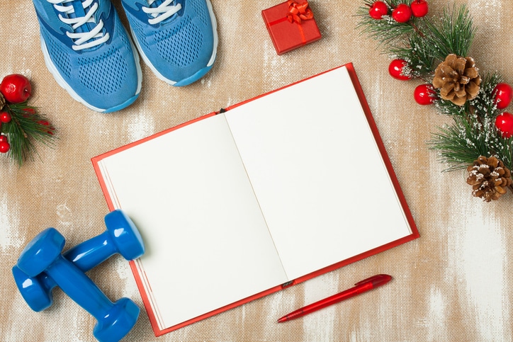 A Red Holiday Fitness Journal Surrounded By Blue Sneakers And Weights, A Red Gift Box And Pen, And A Pine Branch With Red Berries And Cones.