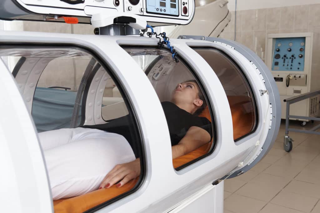 A Young Female Patient In A Black Shirt And White Pants Is Lying In A Hyperbaric Oxygen Therapy Chamber.