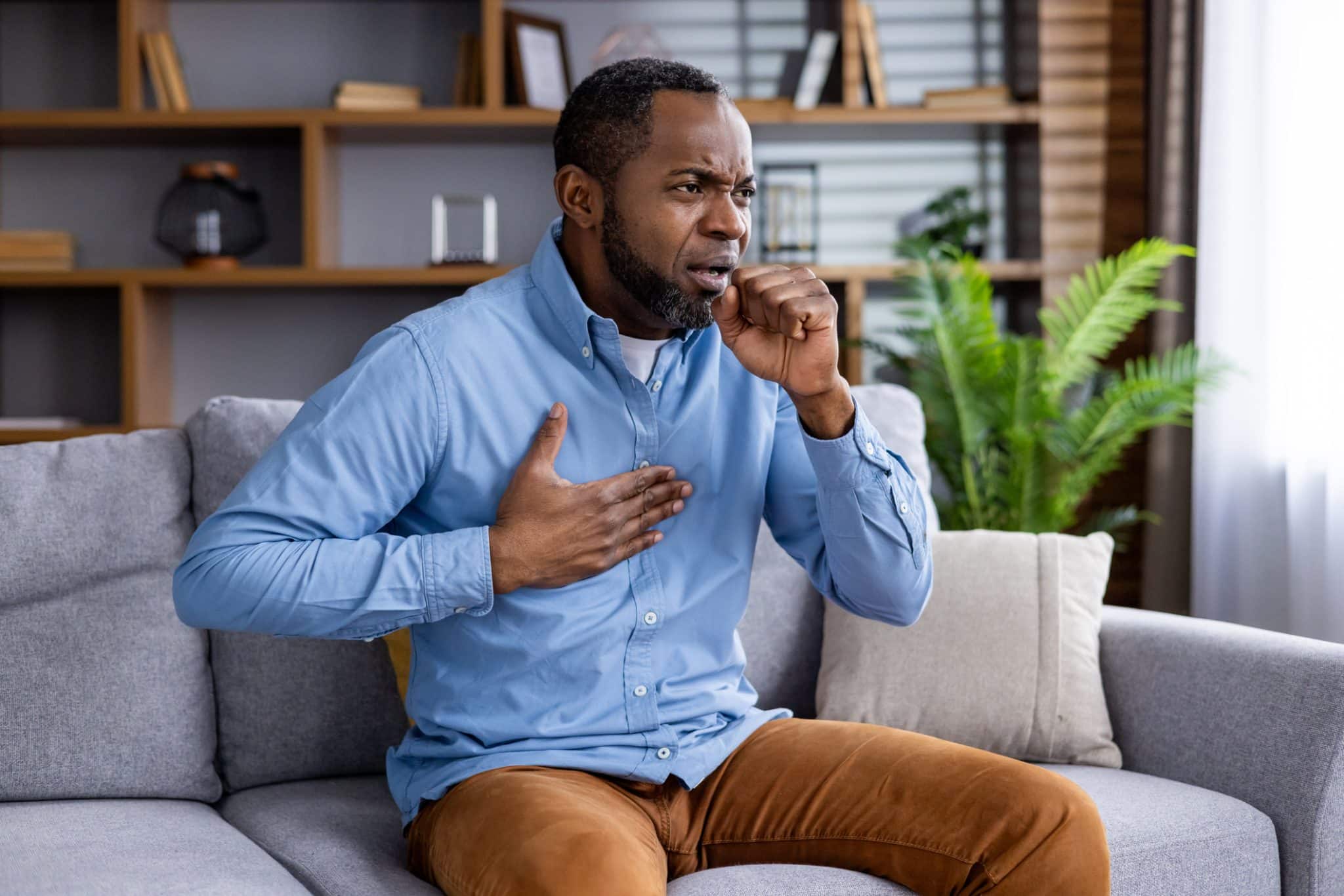 A man sitting on a sofa holding his chest and coughing into his hand while experiencing shortness of breath.