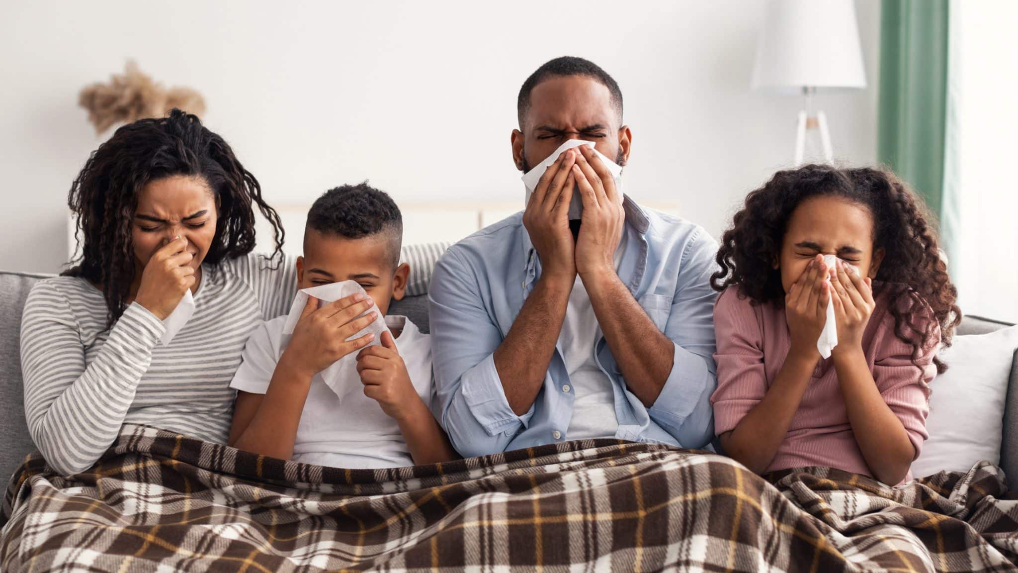 A family suffering from the flu sits together on a couch under a blanket, all blowing their noses.